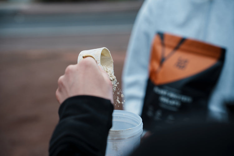 A scoop of plant protein powder tips into a shaker bottle, while a person in the background holds an orange Inna Protein bag.